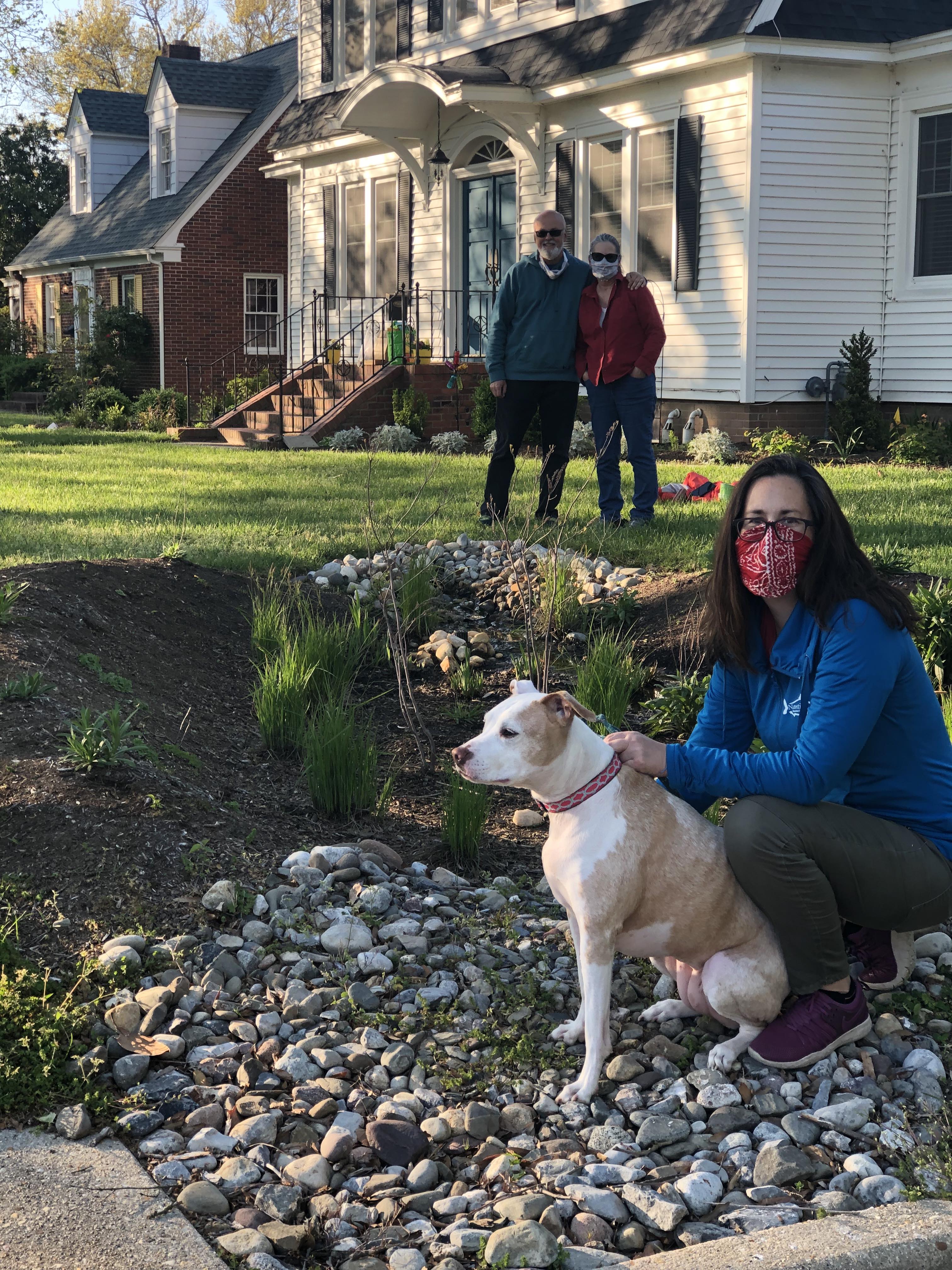 Lisa Wool kneels in the foreground with a medium size brown and white dog at the edge of a rain garden while a couple stands in the background, in their yard in front of a white house