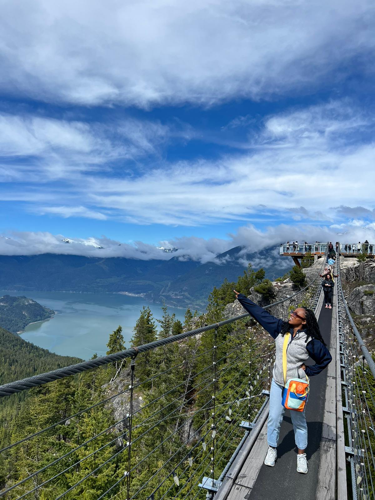 A young woman stands on a walking bridge over a forest naer mountains and a lake. She points toward the sky