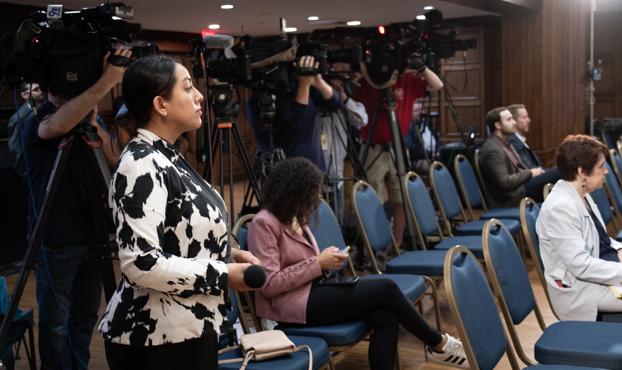 A woman stands with a microphone in front of seated journalists and a row of camera operators.