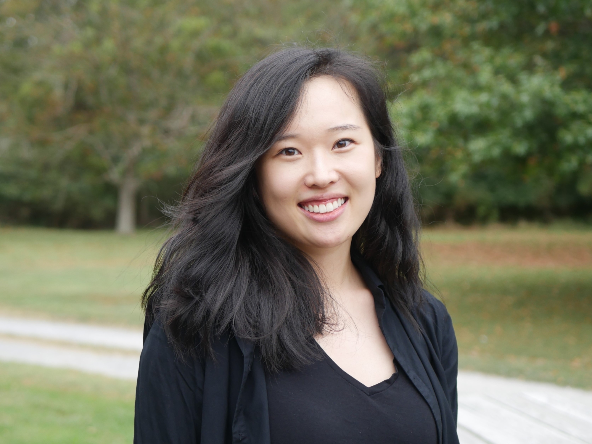 Portrait of Carol Kim outdoors in a grassy area with trees and a path