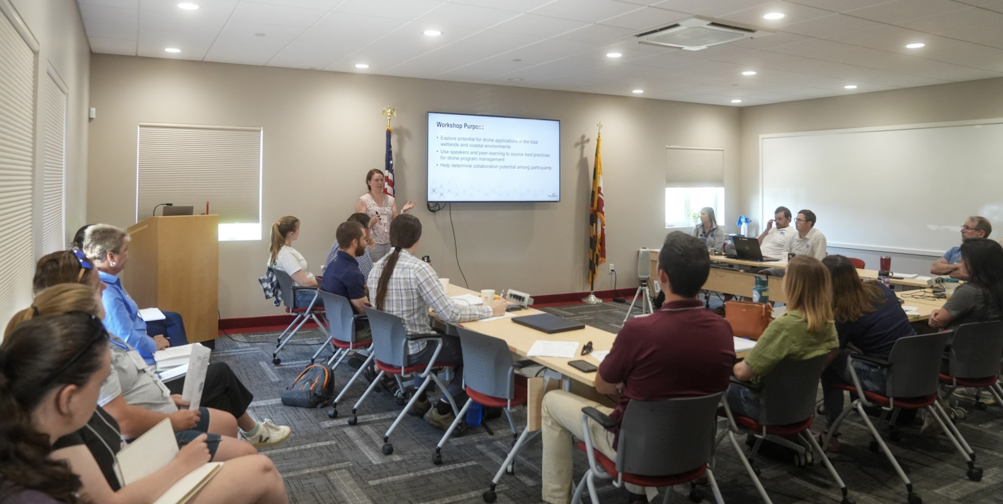 Woman delivers remarks in a conference room to about 20 people sitting around a table and against the walls