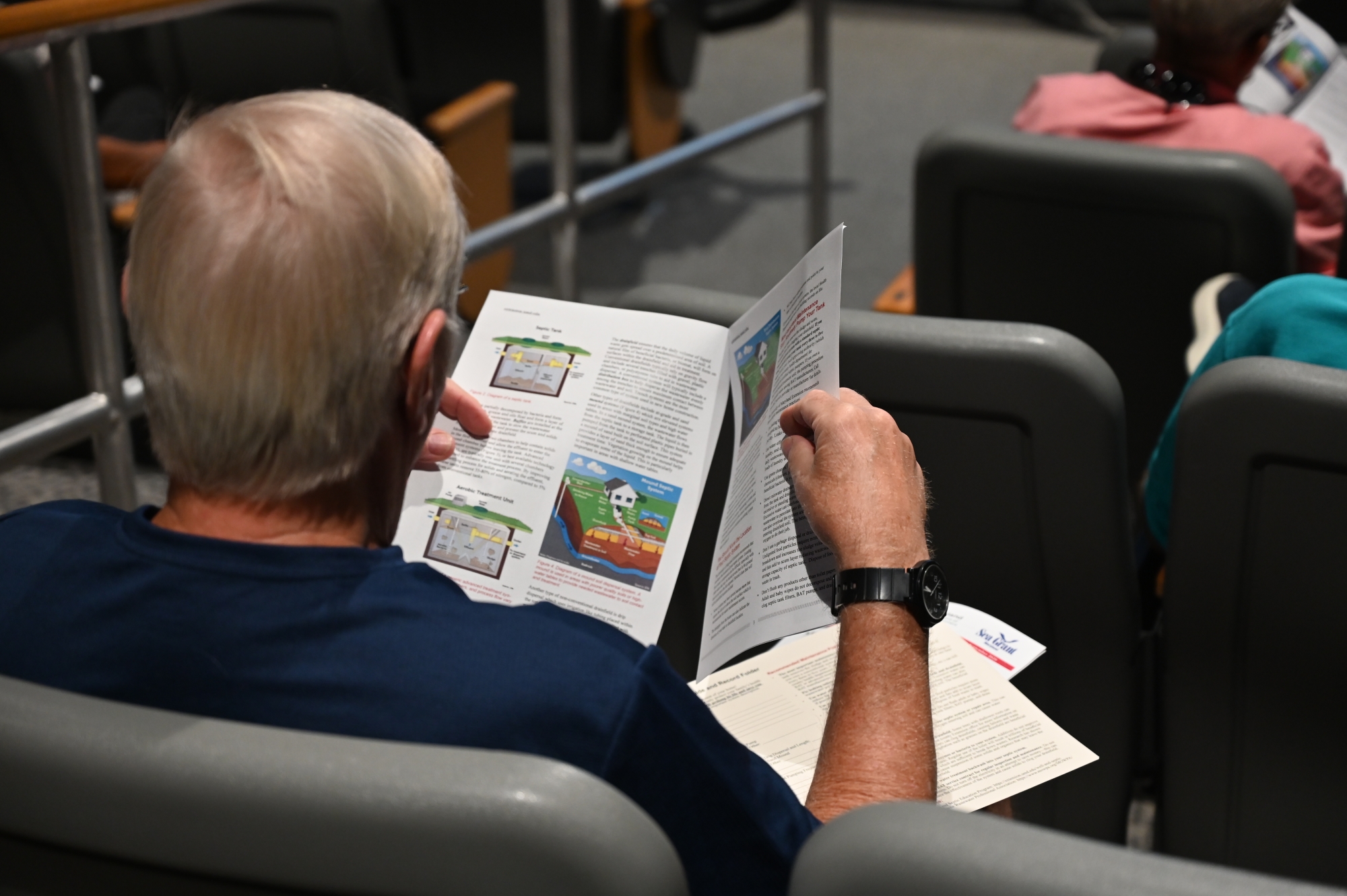 A man seated in an auditorium flips through a packet of papers about septic system care.