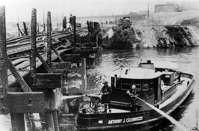 Two firemen on a boat extinguish flames on a river near a railroad bridge.