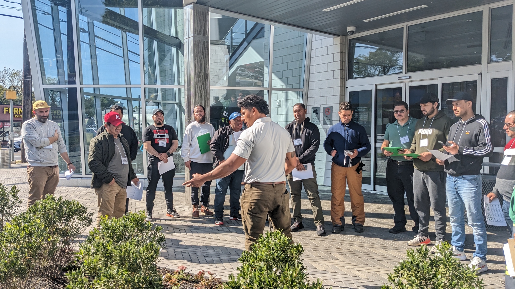 A group of landscaping professionals listen to a speaker demonstrating how to plant trees outside a building with large glass windows