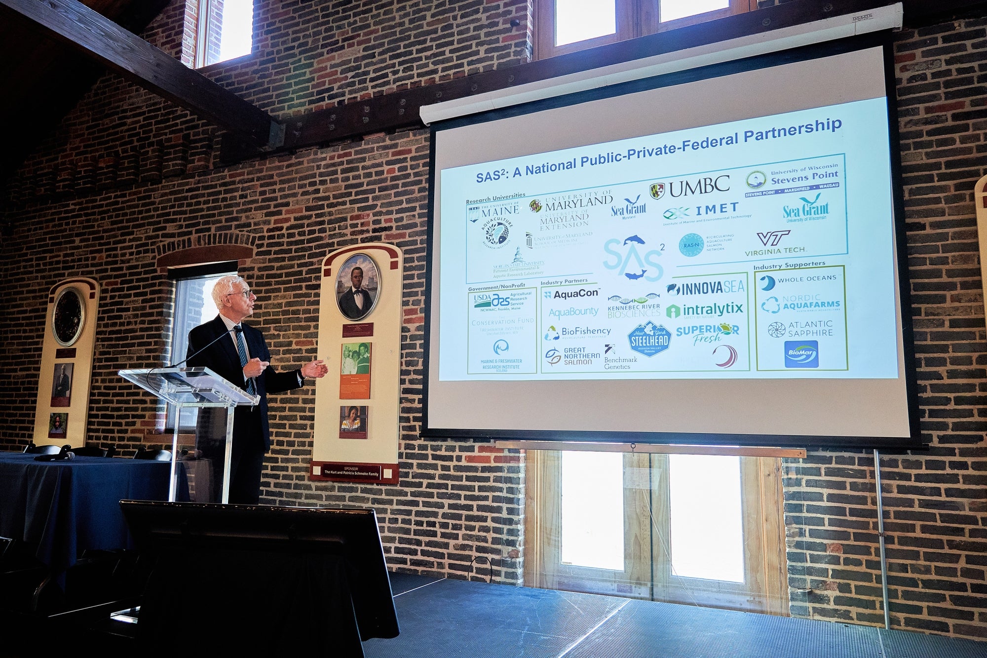 A man standing at a lectern in front of an audience with a powerpoint presentation