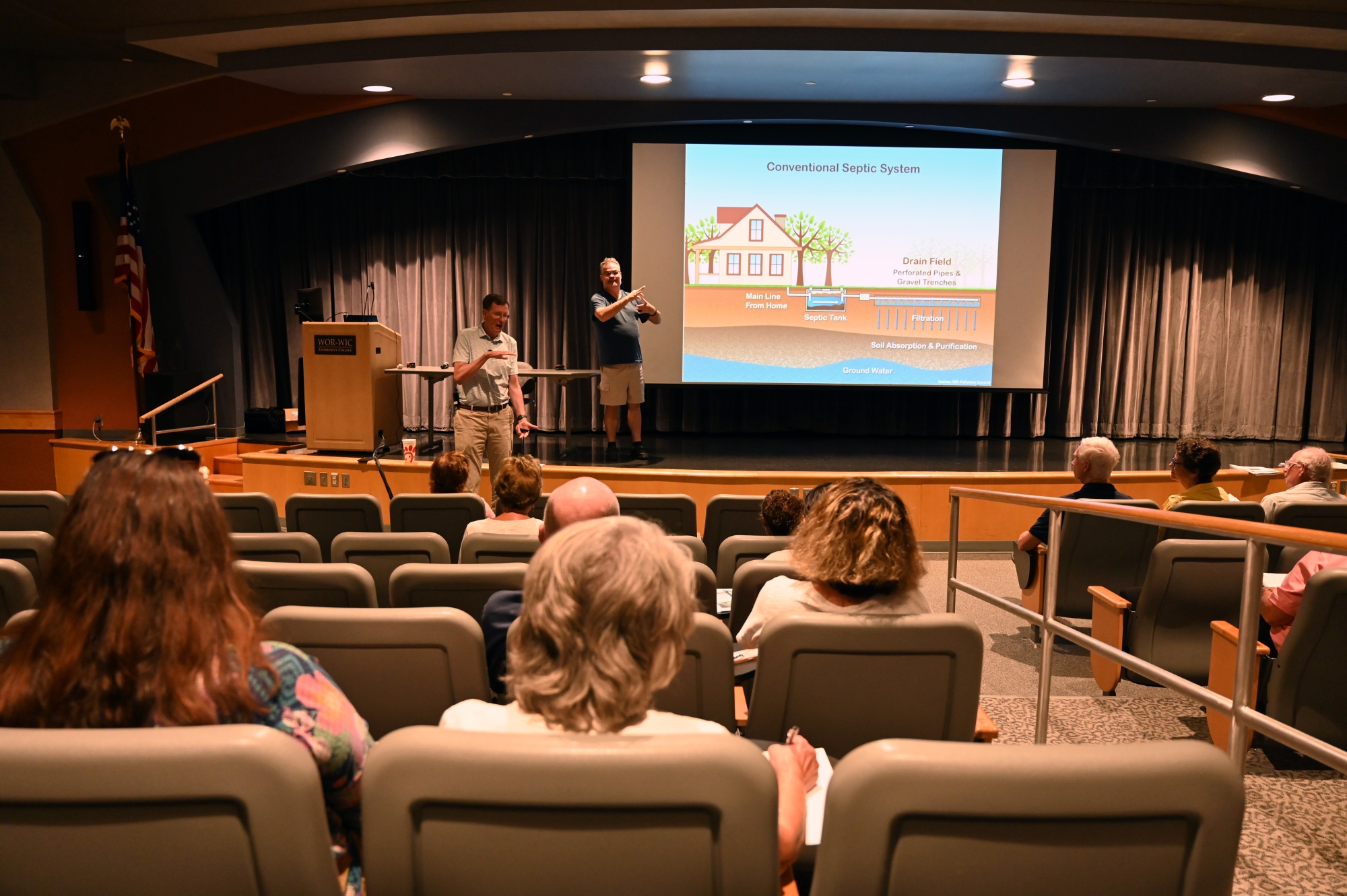 A man and ASL interpreter stand near a stage in front of an auditorium of people.