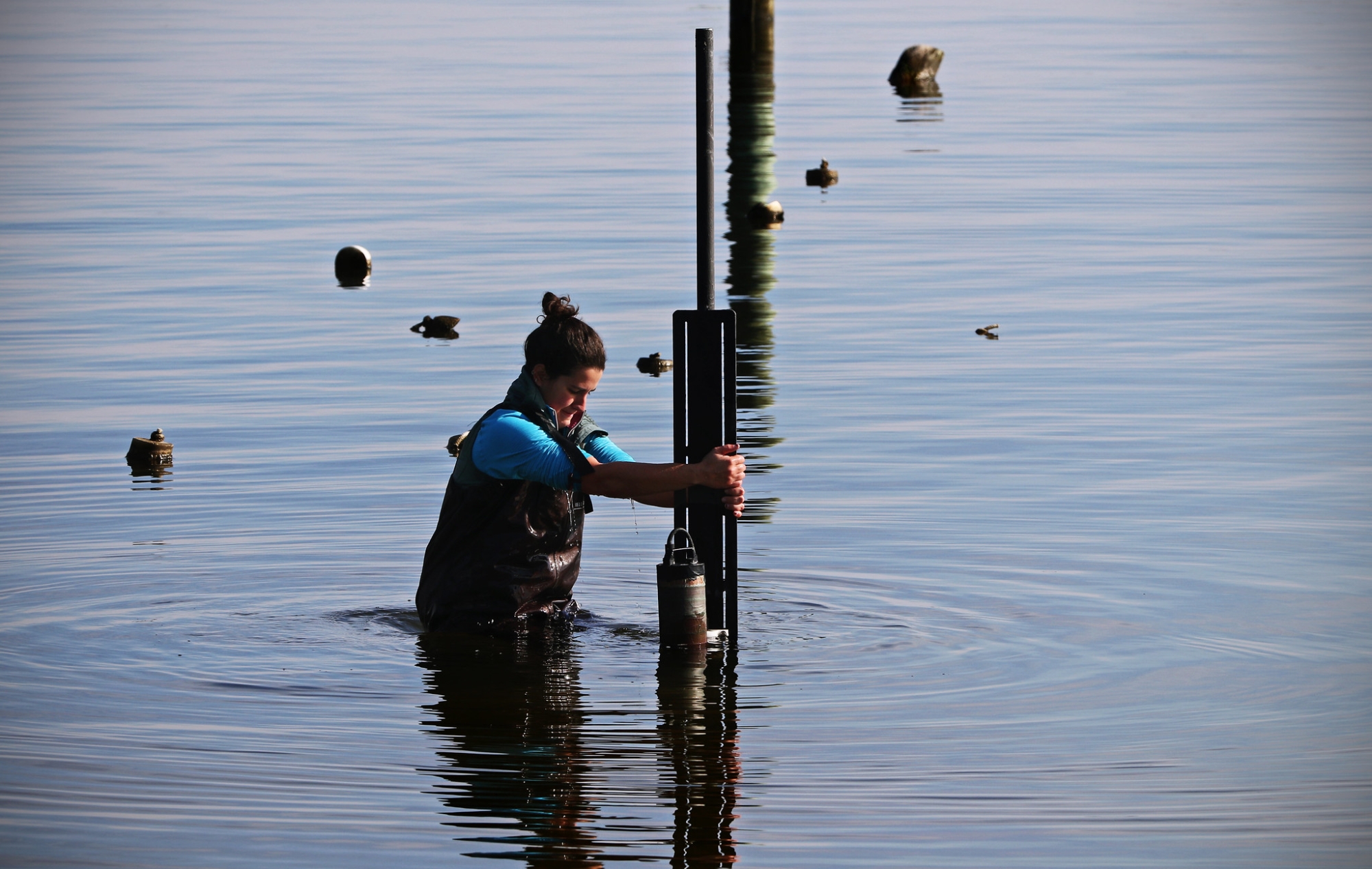 A woman pushes a pole with a sensor attached to it into the water near some rocks and buoys.