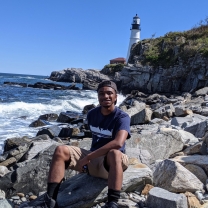 Kai Hardy sits on some rocks near a lighthouse.