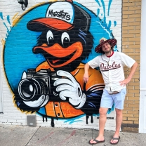 Michael Macon poses next to a mural of the Baltimore Orioles mascot while wearing an Orioles T-shirt, shorts, and a wide-brim hat