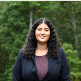 Woman with long brown hair in a black jacket in front of trees