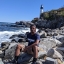 Kai Hardy sits on rocks along a body of water with a lighthouse in the background