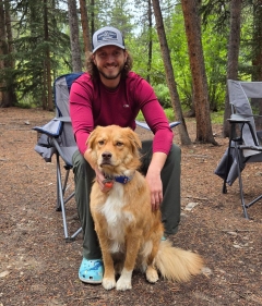 Michael Macon sits in a camp chair and poses with a dog in the woods