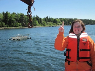 Cat Frederick wearing a PFD standing in front of a body of water, pointing up to a hook overhead